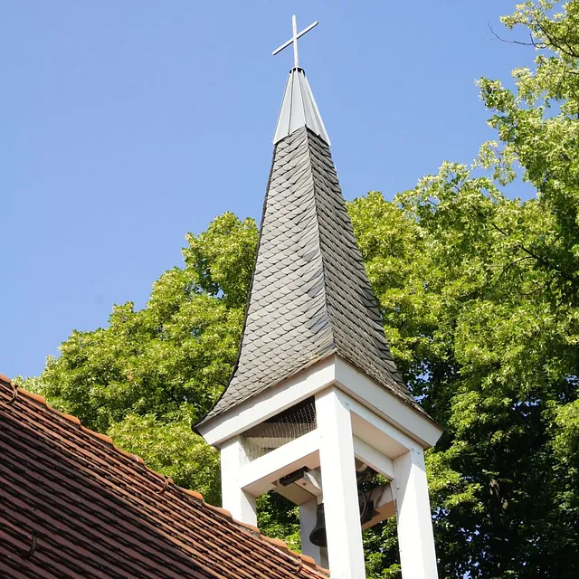 Dachreiter der Gartenstadt-Kirche mit spitzem Helm und Kreuz über dem Ziegeldach, vor blauem Himmel und grünen Baumkronen