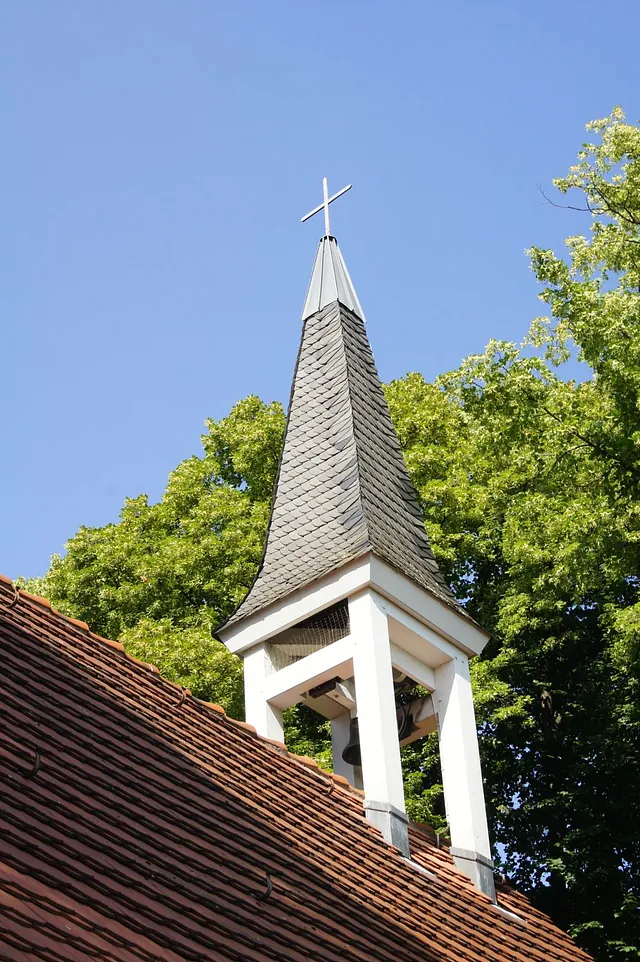 Dachreiter der Gartenstadt-Kirche mit spitzem Helm und Kreuz über dem Ziegeldach, vor blauem Himmel und grünen Baumkronen