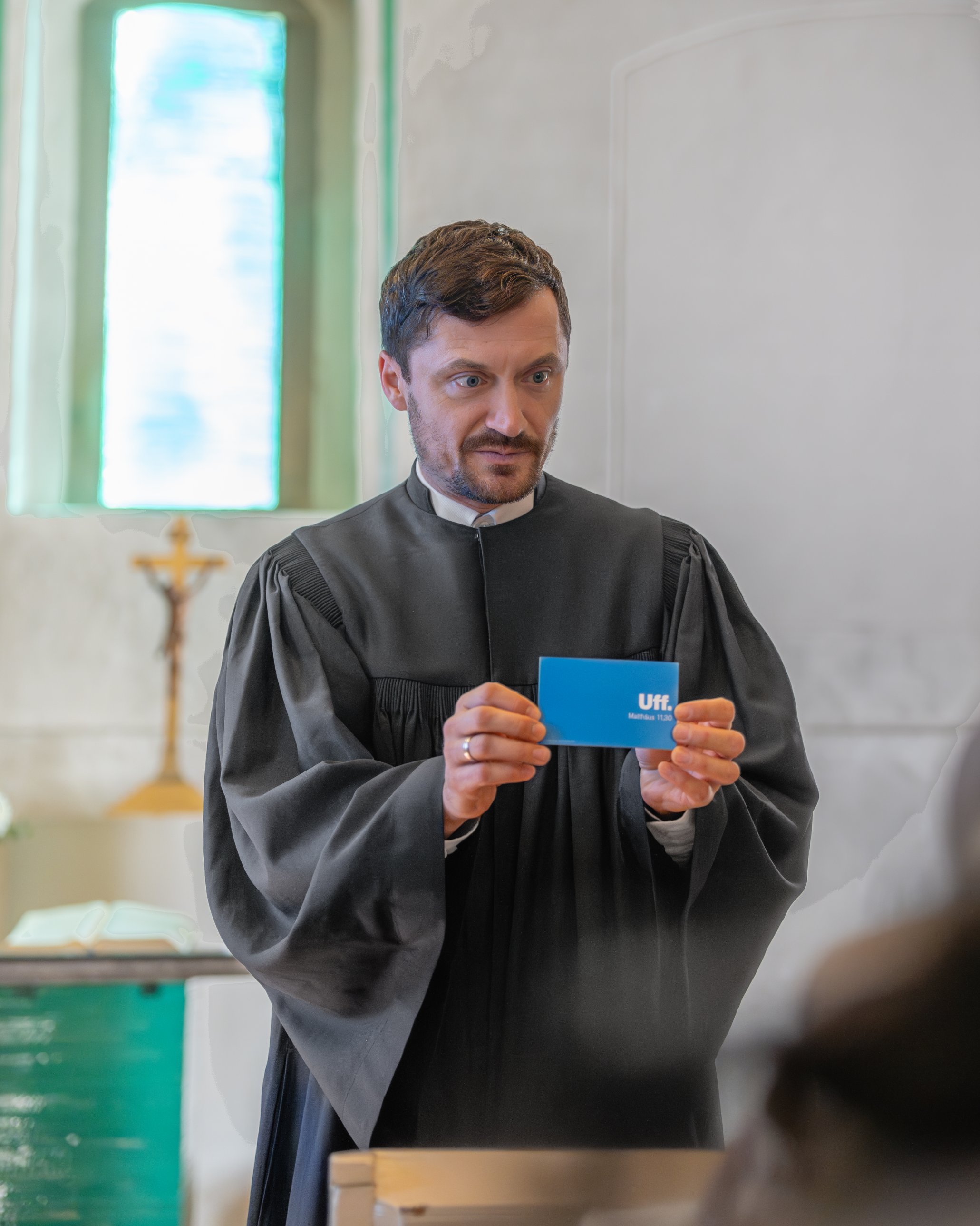 Pfarrer Viktor Weber im Talar hält eine blaue Karte mit der Aufschrift „Uff. Matthäus 11,30“