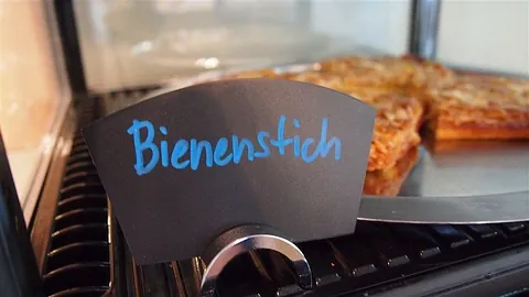 Ein Stück Bienenstich in der Kuchenvitrine des Café Pi8, davor ein Schild mit der Aufschrift Bienenstich