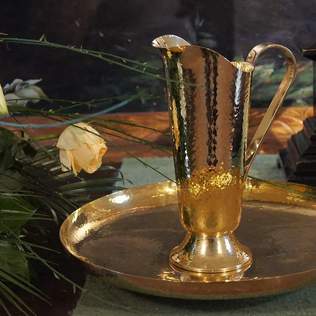 Taufschale aus gehämmertem Messing mit passender goldfarbener Taufkanne zwischen weißen Lilien