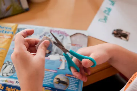 Kinderhände schneiden mit einer türkisen Schere ein Bild aus einer Zeitschrift aus