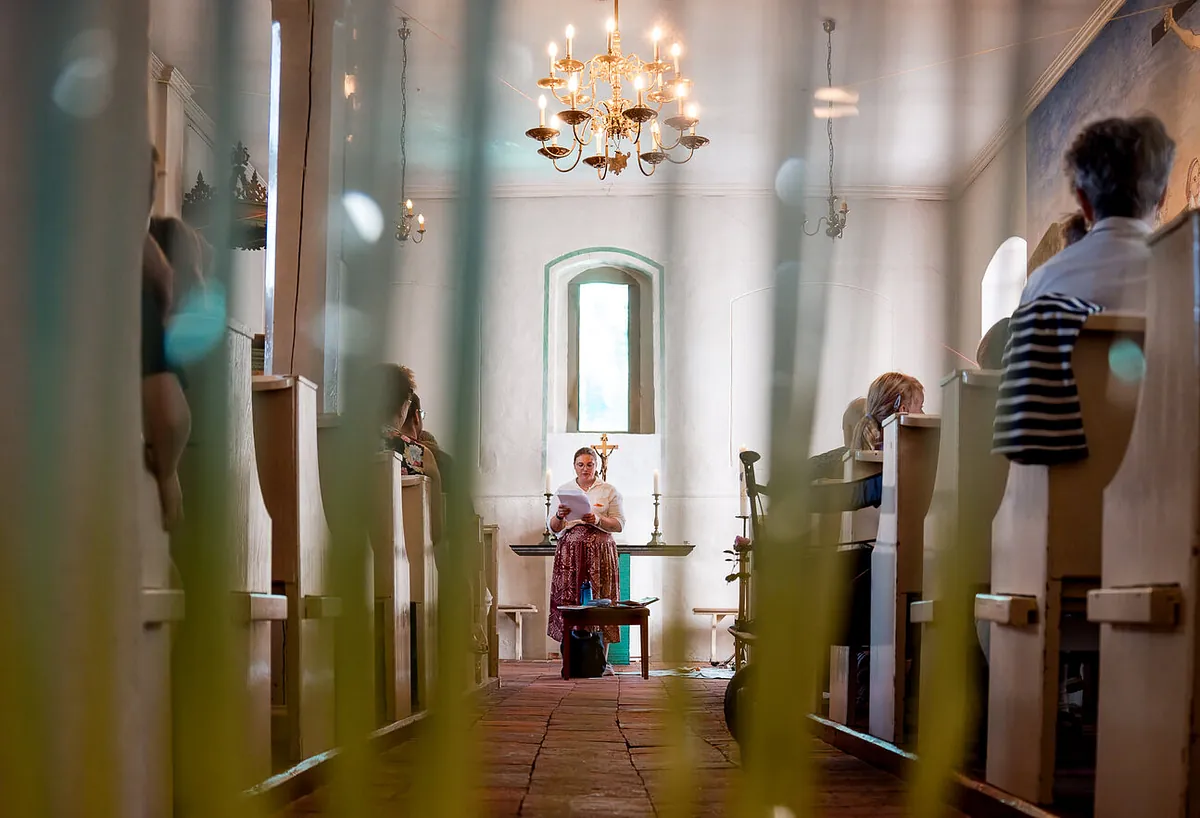 Blick durch hohe Gräser in den Mittelgang der Dorfkirche Alt-Staaken: Eine Frau liest am Altar, Menschen sitzen in den Bänken, darüber hängt ein Kronleuchter mit Kerzen