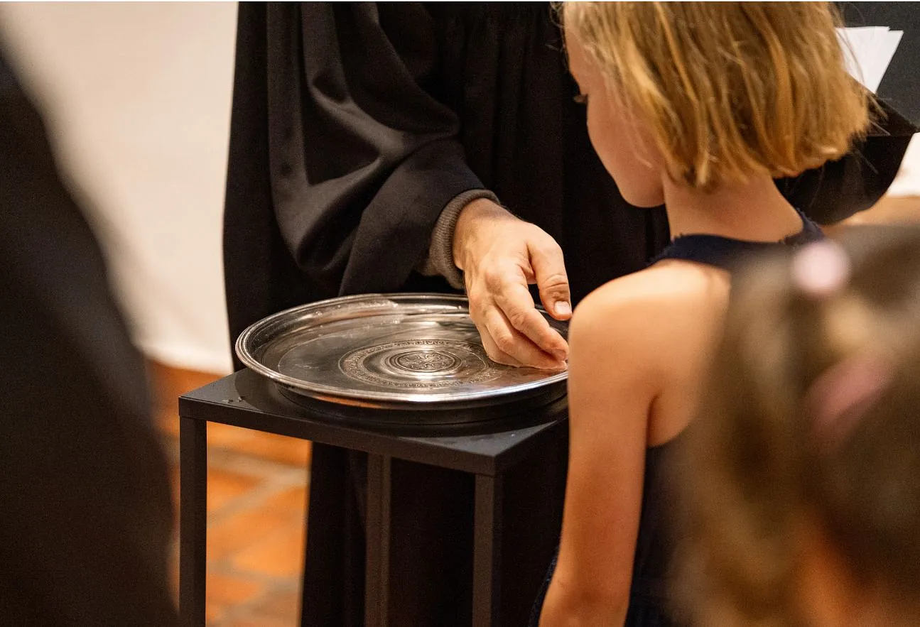 Hand schöpft Wasser aus dem Taufbecken, daneben steht ein Mädchen