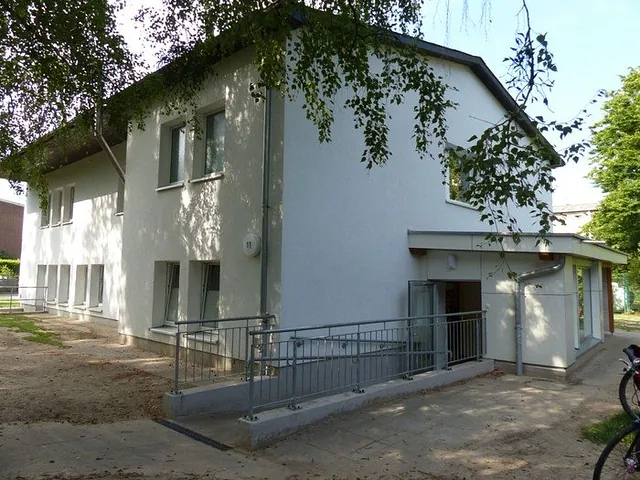 Zweigeschossiges weißes Kitagebäude der Kita Arche Noah mit Rampe zum Eingang, davor Bäume
