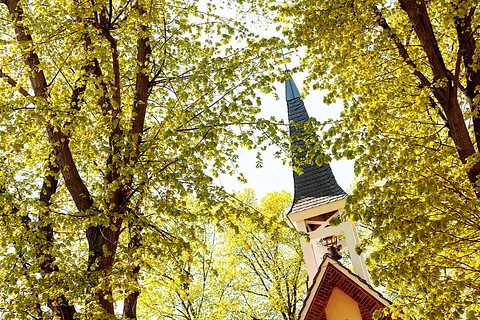 Kirche Staaken-Gartenstadt: schiefergedeckter Turmhelm mit offenem Glockenstuhl über rotem Backsteingiebel, eingerahmt von frisch grünen Baumkronen