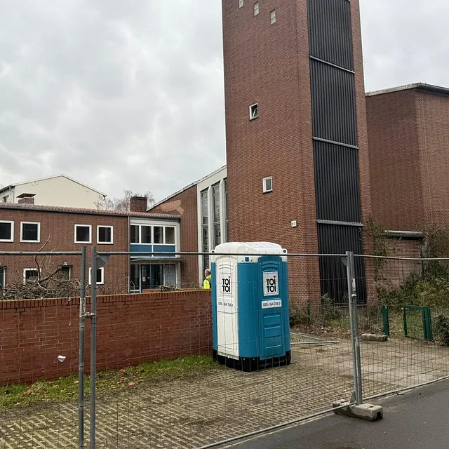 Backsteinturm der ehemaligen Zuversichtskirche mit dunklen Fensterbändern neben dem Kirchbau, davor Bauzaun und Baustellentoilette