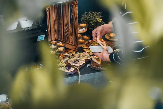 Hände nehmen Fingerfood von einem Buffet mit Holzbrettern und kleinen Gläsern, im Vordergrund unscharfe Blätter