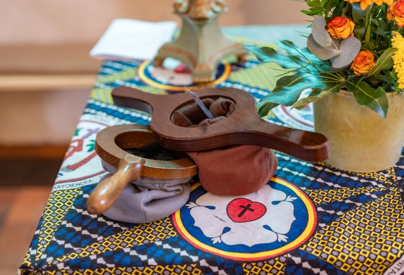 Zwei Kollektenbeutel liegen auf einem bunt gemusterten Altartuch; rechts steht ein Blumenstrauß, im Hintergrund sind weitere Gegenstände im Kirchenraum unscharf zu sehen.