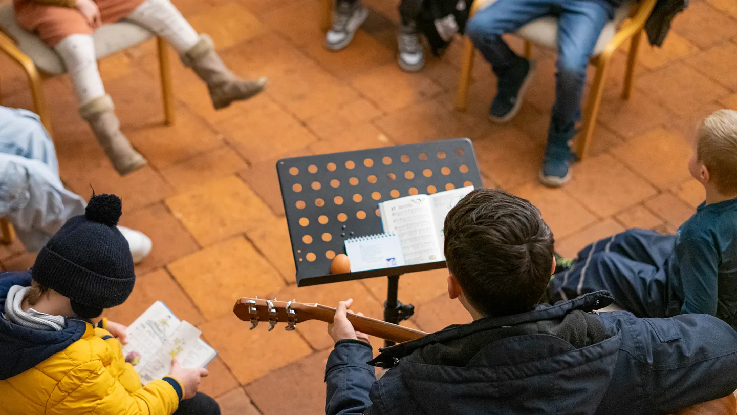 Menschen sitzen im Kreis auf einem Holzboden, ein junger Mann spielt Gitarre vor einem Notenständer