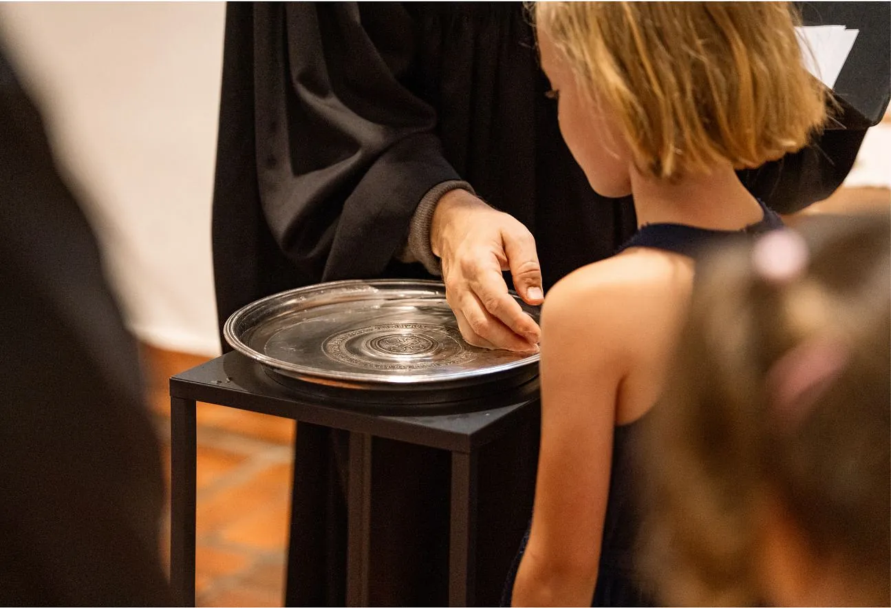 Eine Hand schöpft Wasser auf dem Taufbecken. Ein KInd steht daneben
