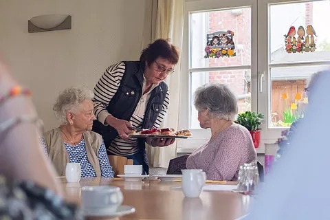 Eine Ehrenamtliche reicht Seniorinnen an einer Kaffeetafel ein Tablett mit Kuchen