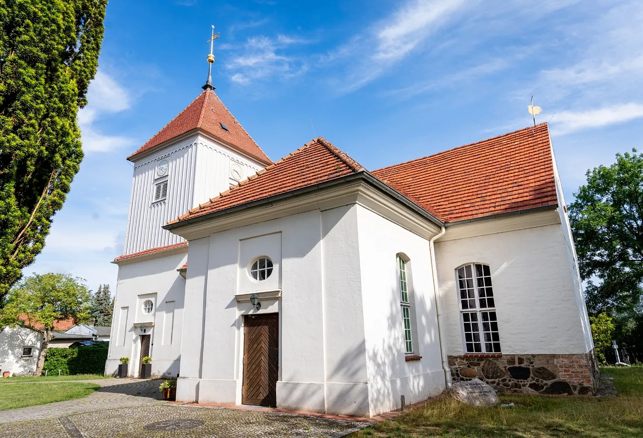 Weiße Dorfkirche Alt-Staaken mit rotem Ziegeldach und Kirchturm vor blauem Himmel. Seitlich sind der Eingang, hohe Kirchenfenster sowie Bäume und Grünflächen zu sehen.