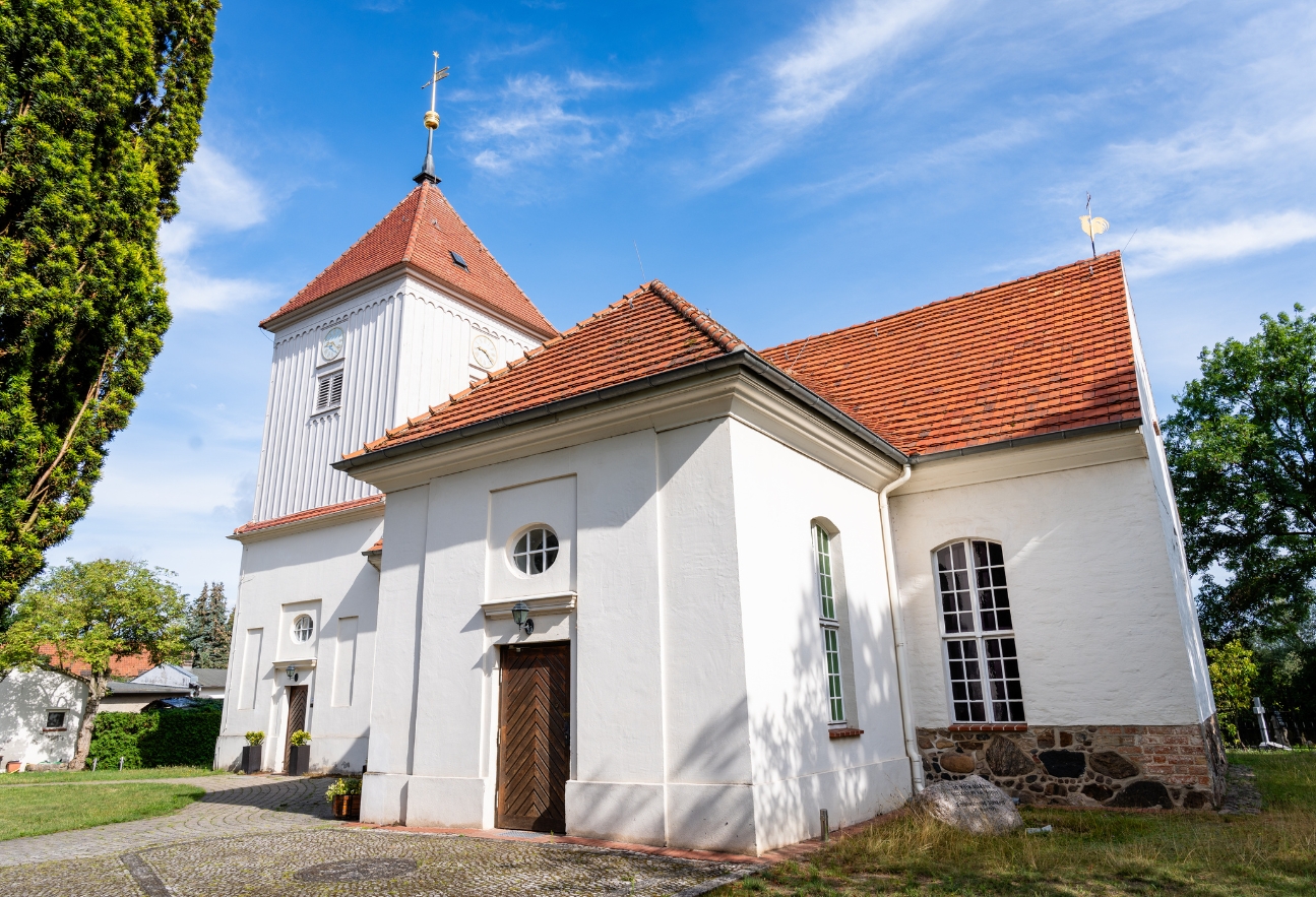 Weiße Dorfkirche Alt-Staaken mit rotem Ziegeldach und Kirchturm vor blauem Himmel. Seitlich sind der Eingang, hohe Kirchenfenster sowie Bäume und Grünflächen zu sehen.