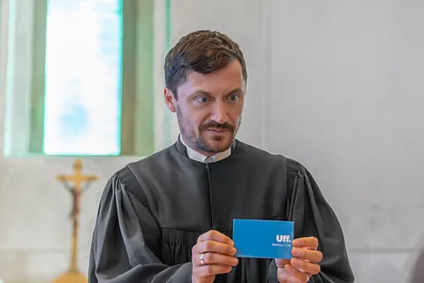 Pfarrer Viktor Weber im Talar hält eine blaue Karte mit der Aufschrift „Uff. Matthäus 11,30“