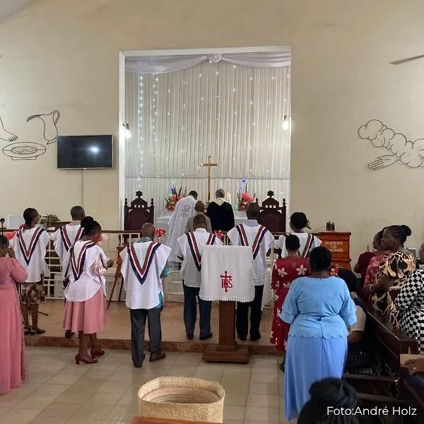 Menschen stehen in einer Kirche vor dem Altar; einige tragen weiße Chorumhänge, im Vordergrund und auf den Bänken ist die Gemeinde zu sehen.
