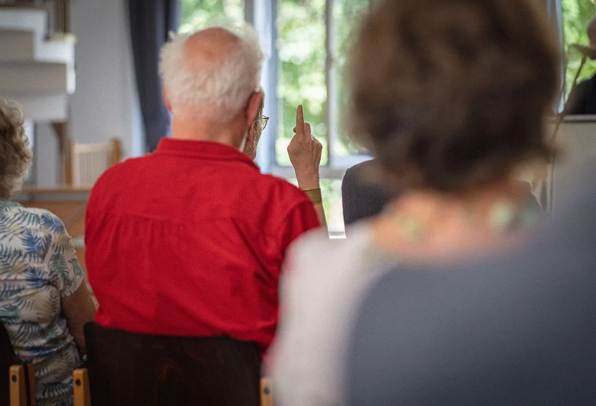 Menschen sitzen in einem hellen Raum auf Stühlen; in der Mitte ist eine Person in rotem Hemd von hinten zu sehen, im Hintergrund hebt jemand einen Finger zum Reden.