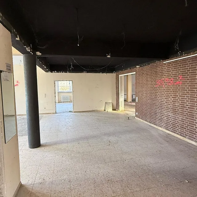 Leergeräumter Saal im alten Gemeindehaus: offene Decke, schwarze Stütze, Backsteinwand und Blick durch eine Tür in den Flur