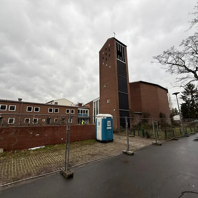 Das alte Gebäudeensemble der Zuversichtskirche mit Glockenturm, Kirchbau und Gemeindehaus hinter einem Bauzaun am Gehweg