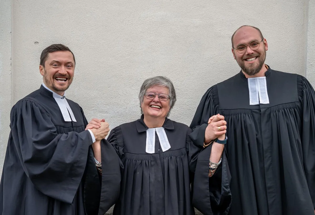 3 Pfarrpersonen im Talar halten sich an den Händen und lächeln