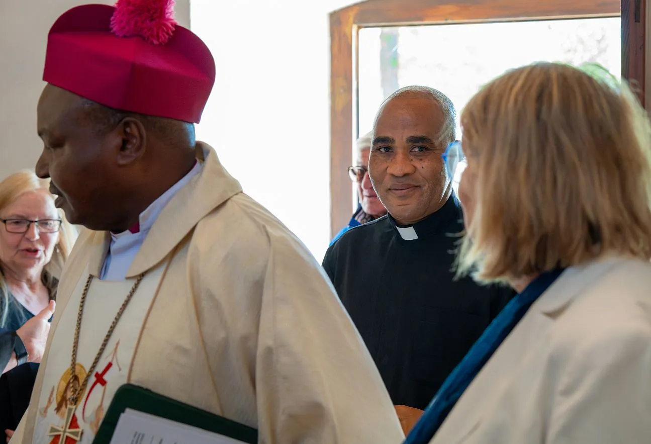 Mehrere Menschen stehen in einem hellen Raum beieinander; links ist ein Geistlicher in liturgischer Kleidung mit roter Kopfbedeckung zu sehen, daneben weitere Personen im Gespräch.