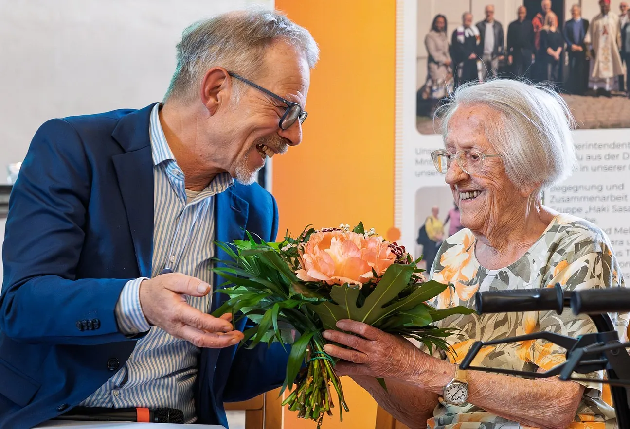 Ein Mann im blauen Sakko überreicht einer älteren Dame einen Blumenstrauß, beide lachen sich herzlich an