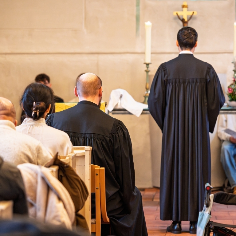 Blick in einen Gottesdienst: Zwei Personen im schwarzen Talar stehen bzw. sitzen vor dem Altar, während die Gemeinde in den Kirchenbänken Platz genommen hat. Im Hintergrund sind Kerzen und ein Kreuz zu sehen.