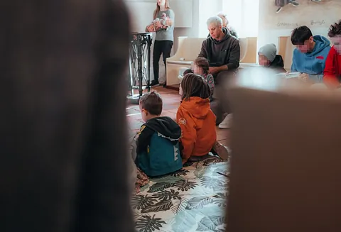 Kinder sitzen von hinten gesehen auf einem Teppich im Gottesdienst, dahinter Erwachsene