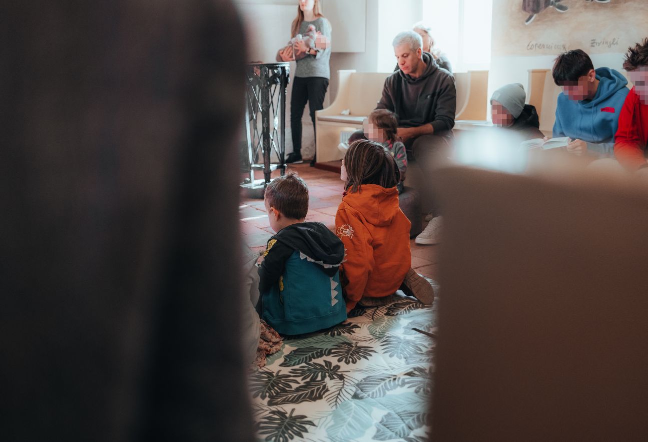 Kinder sitzen von hinten gesehen auf einem Teppich im Gottesdienst, dahinter Erwachsene