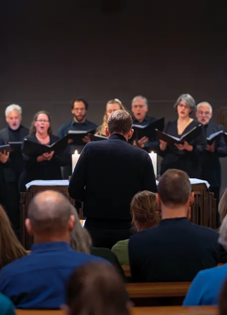 Ein Kirchenchor singt während eines Gottesdienstes; ein Chorleiter steht vor den Sängerinnen und Sängern, im Vordergrund sitzt die Gemeinde in den Kirchenbänken.