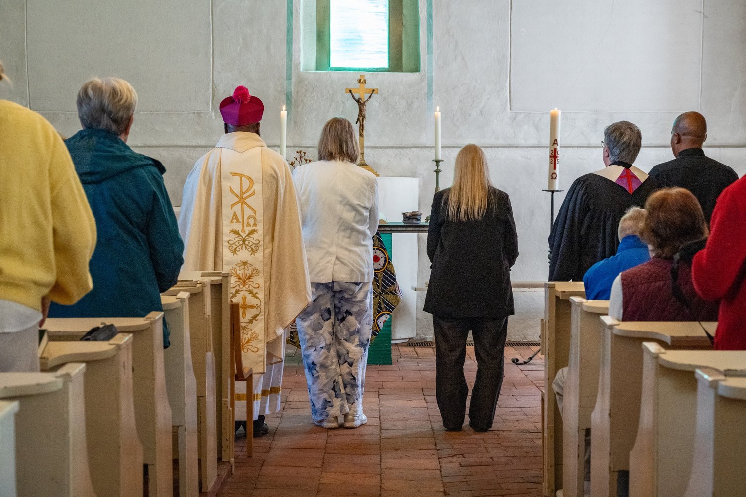 Gottesdienst in der Dorfkirche Alt-Staaken, von hinten gesehen: Geistliche in Gewändern und Gäste stehen gemeinsam vor dem Altar mit Kerzen und Kruzifix