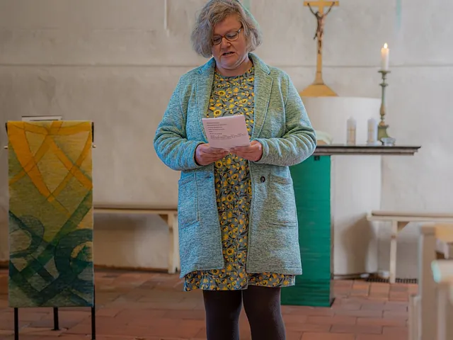 Eine Frau in hellgrüner Jacke liest in einer Dorfkirche vor dem Altar mit Kreuz und Kerze von einem Blatt vor