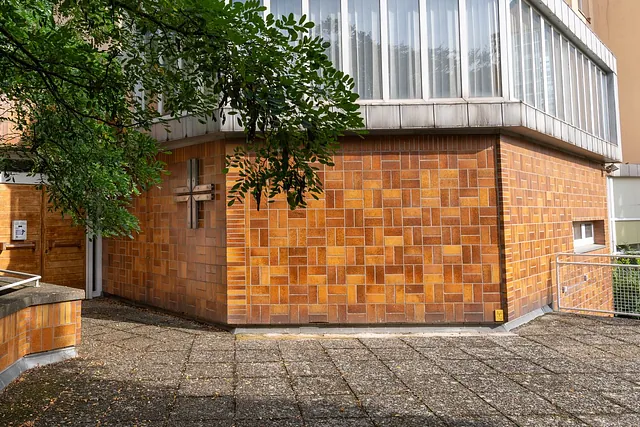 Der vieleckige Kirchraum von außen mit Kreuz an der Fliesenfassade, darüber die Fensterfront des Gemeinwesenzentrums