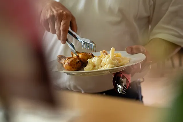 Ein Mann im weißen Poloshirt legt mit einer Zange Hähnchenkeulen und Kartoffelsalat auf einen Teller