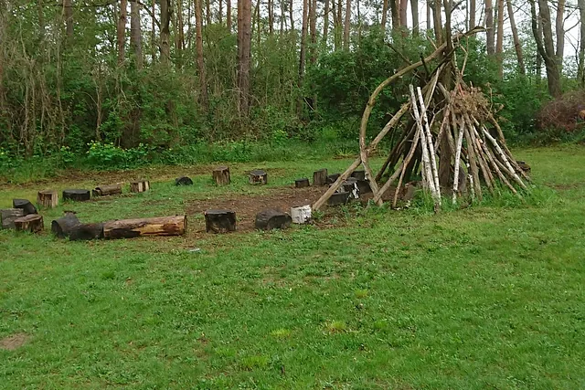Feuerplatz auf der Wiese am Waldrand mit Sitzkreis aus Baumstämmen und einem Tipi aus Ästen